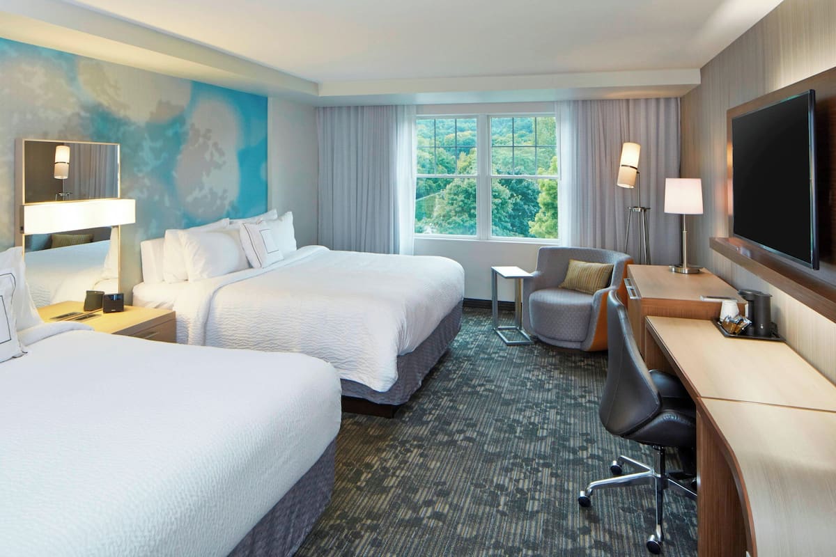 A hotel room with two beds, a desk, a chair, a TV, and a window with a view of trees.
