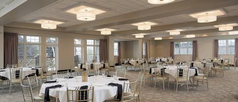 A spacious banquet hall with multiple round tables set for dining, large windows, and a high ceiling with recessed lighting.