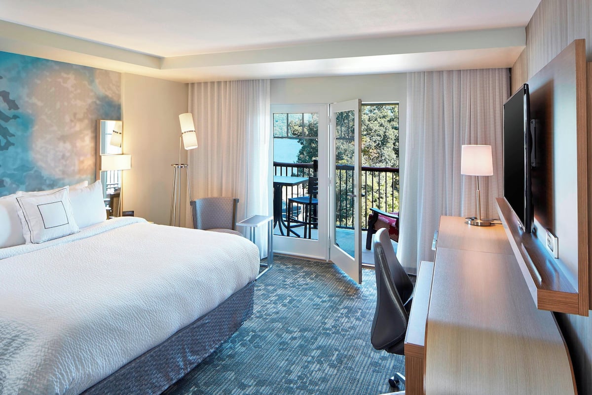 A hotel room with a large bed, a desk with a TV, a balcony with a table and chairs, and a painting on the wall.