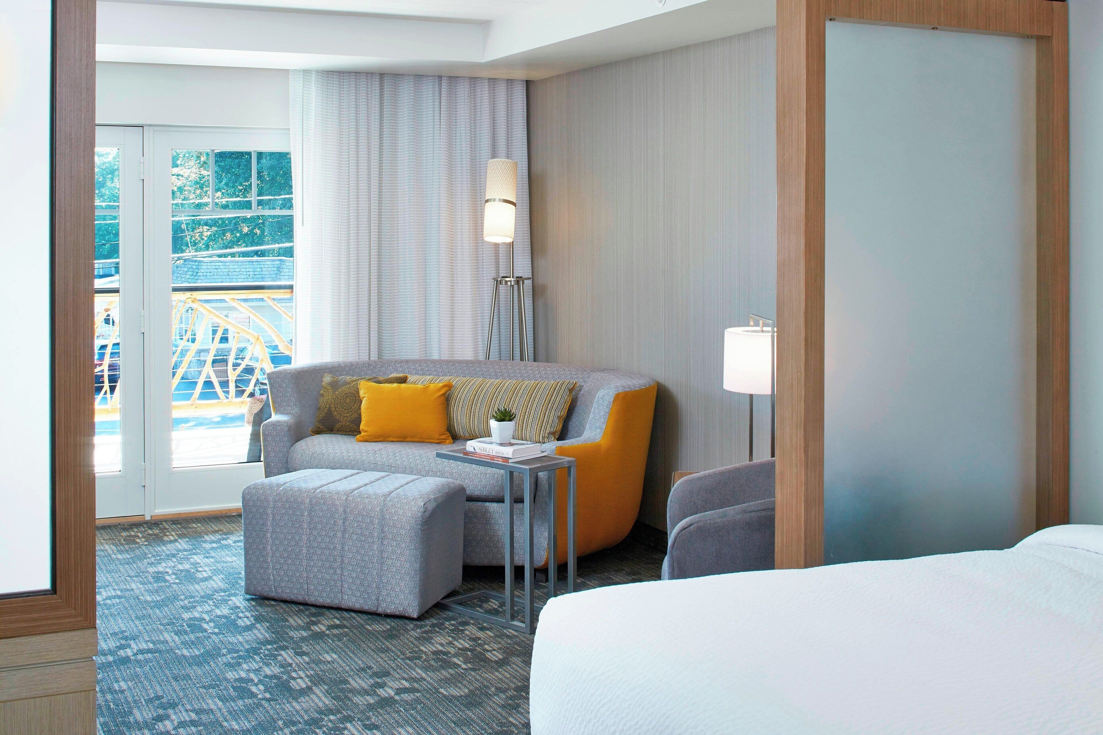 A hotel room with a grey sofa, ottoman, and a bed. There is a lamp, a small table, and a view of a bridge outside.