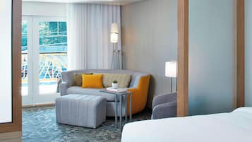 A hotel room with a grey sofa, ottoman, and a bed. There is a lamp, a small table, and a view of a bridge outside.