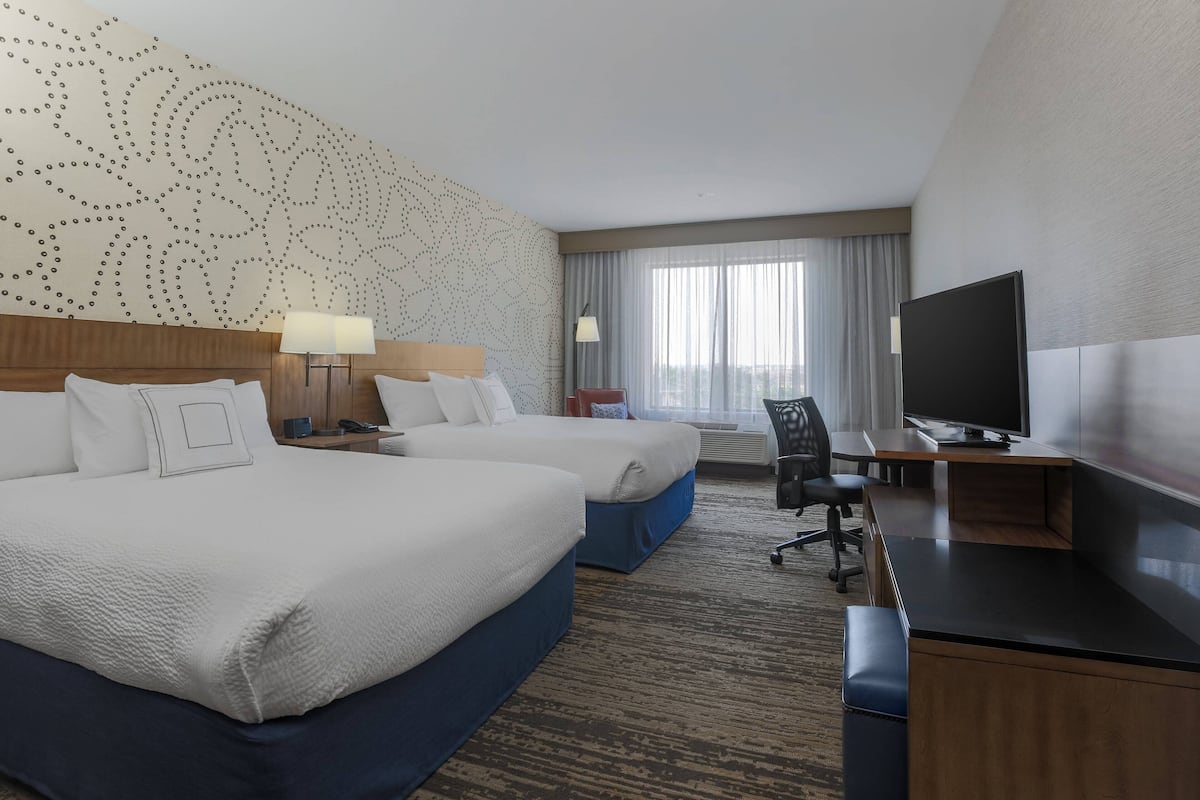 A hotel room with two beds, a desk, a TV, and a large window with curtains.