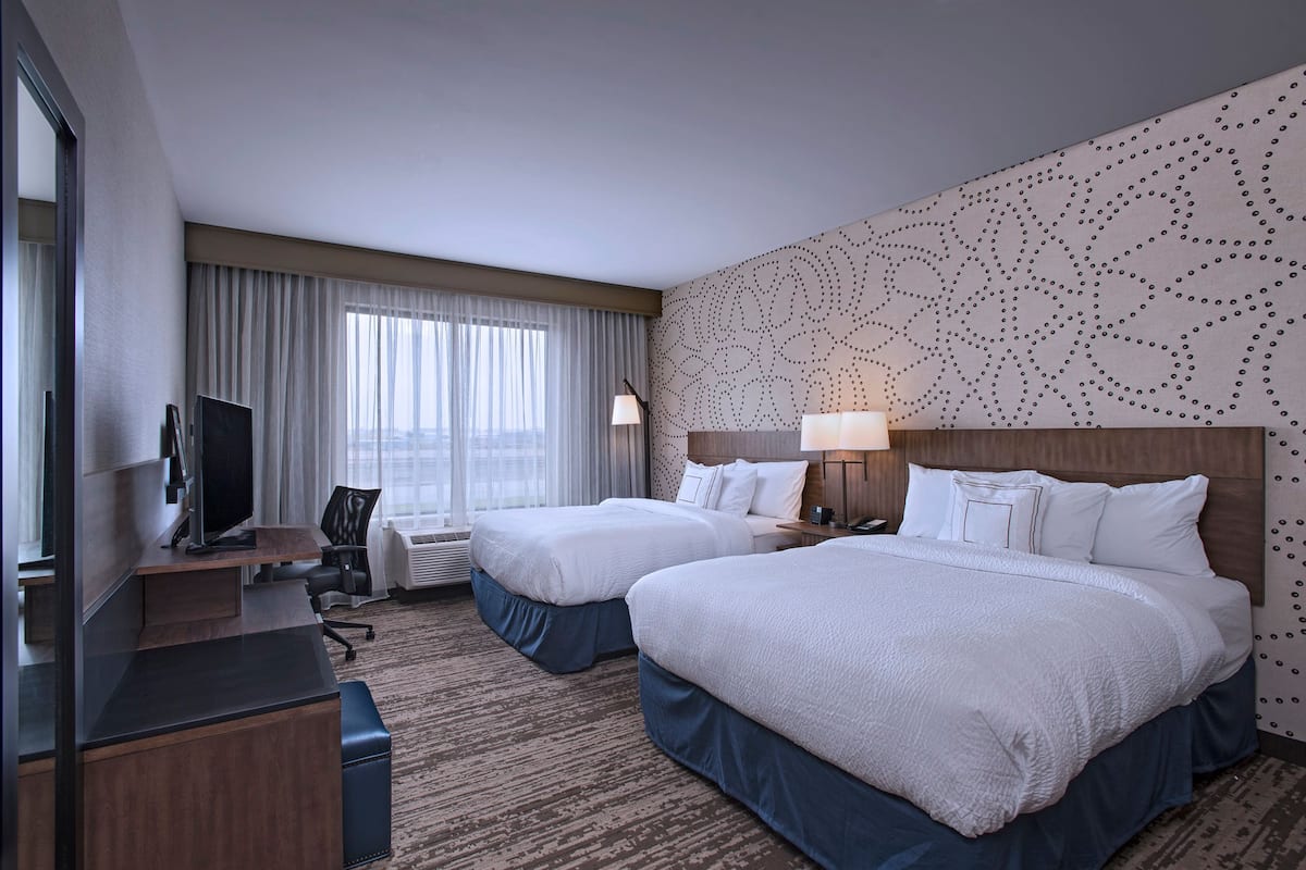 A hotel room with two beds, a desk, a TV, and a large window with curtains.