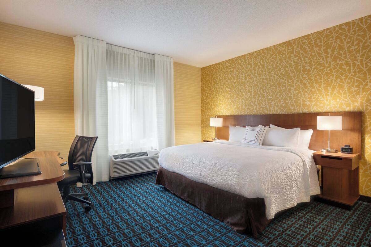 A hotel room with a bed, a desk with a TV, a chair, a lamp, and a patterned carpet.