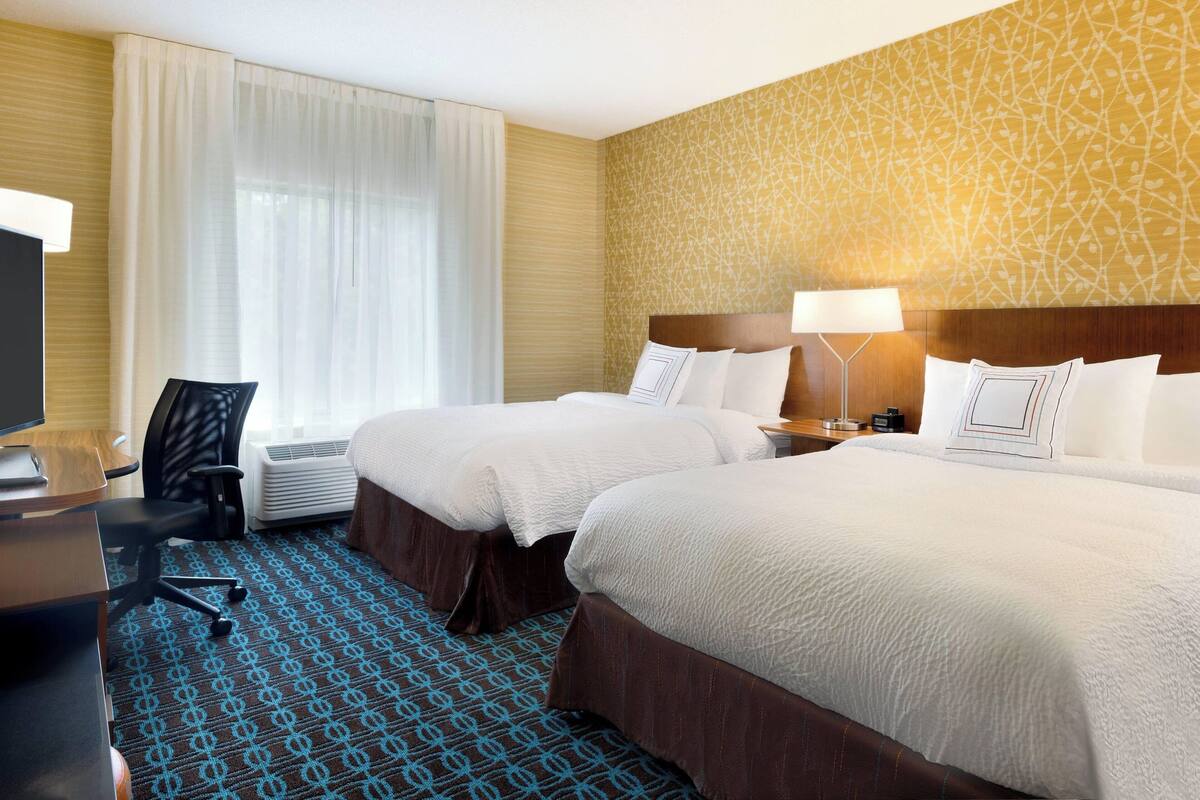A hotel room with two beds, a desk with a chair, a TV, and a lamp.