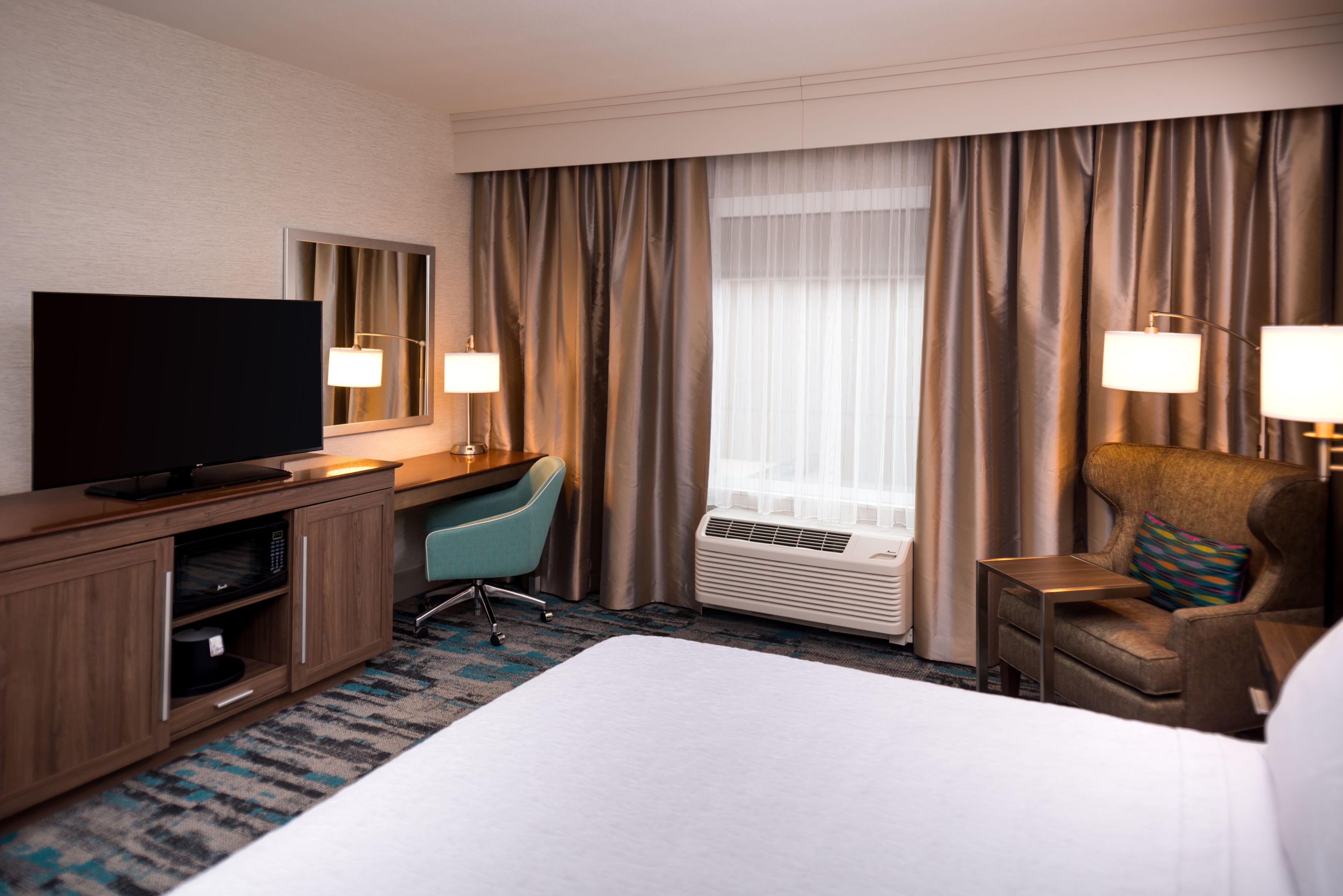 Une chambre d’hôtel comprenant un lit, un bureau avec un téléviseur à écran plat, une chaise, une lampe de bureau, un appareil de climatisation et une fenêtre avec des rideaux.