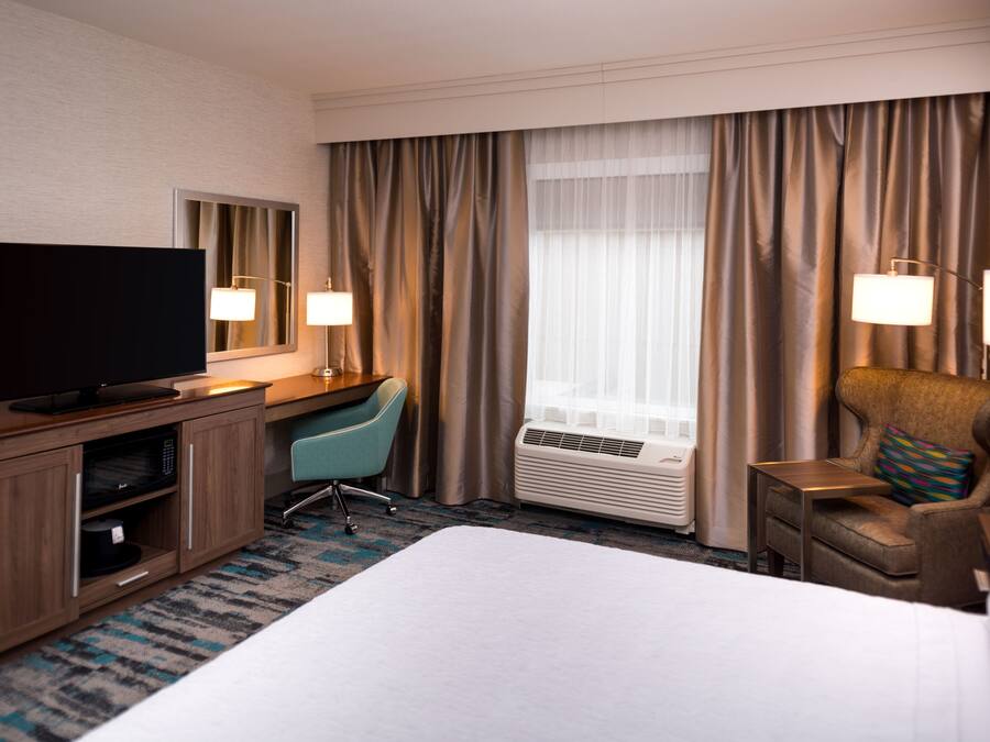 Ein Hotelzimmer mit Bett, Schreibtisch mit Flachbildfernseher, Stuhl, Schreibtischlampe, Klimaanlage und einem Fenster mit Vorhängen.