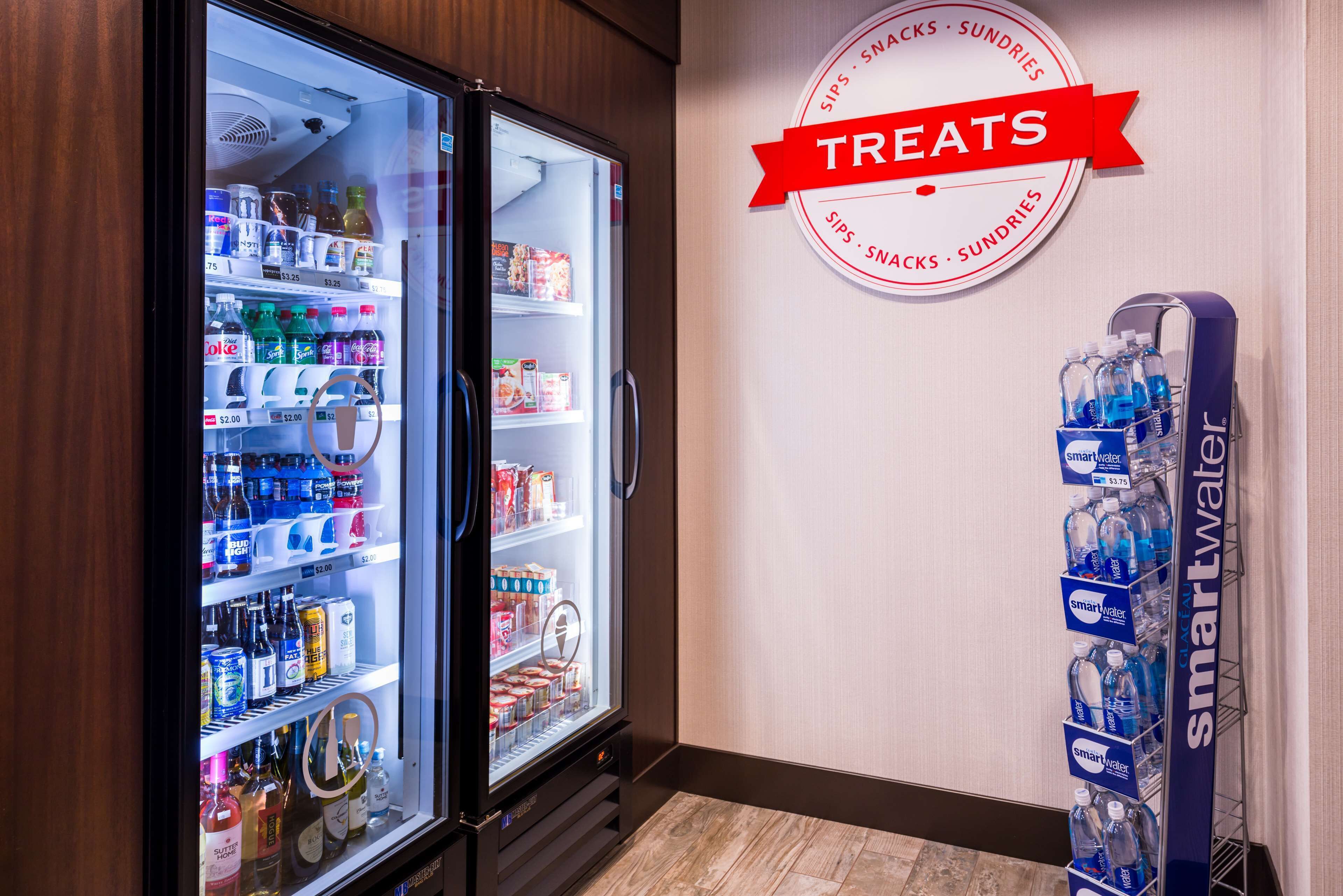 Máquina expendedora con diversas bebidas y aperitivos, un refrigerador lleno de bebidas y alimentos, y un cartel publicitando golosinas.