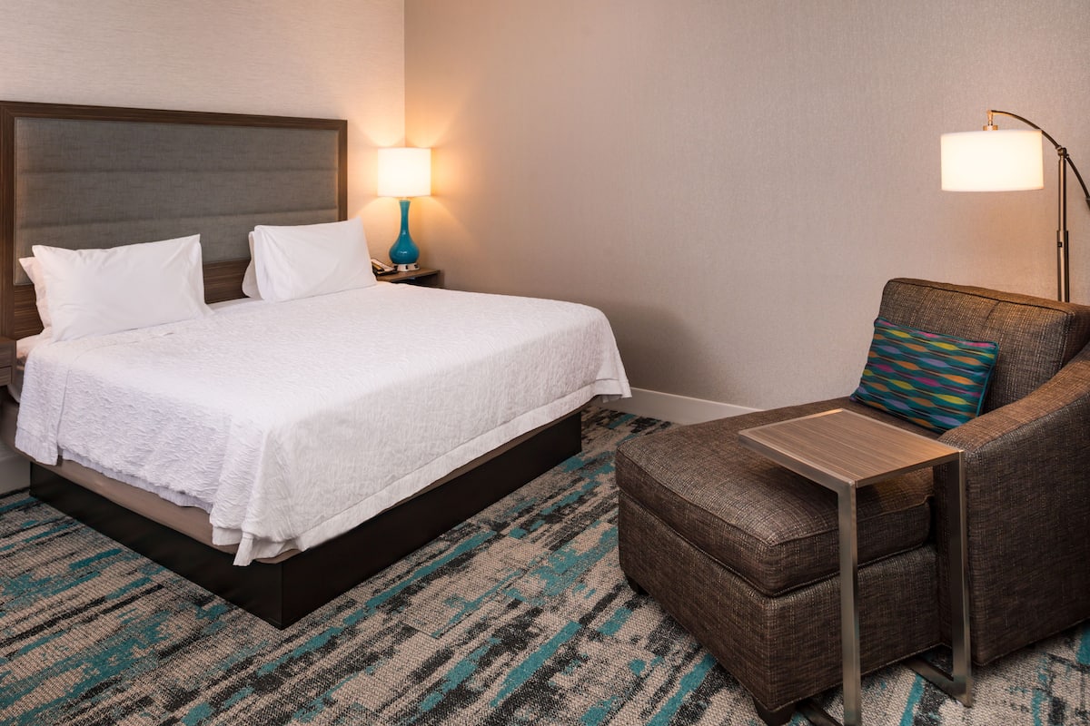 Ein Hotelzimmer mit einem Bett, einem Nachttisch mit Lampe, einem Sessel und einem gemusterten Teppich.