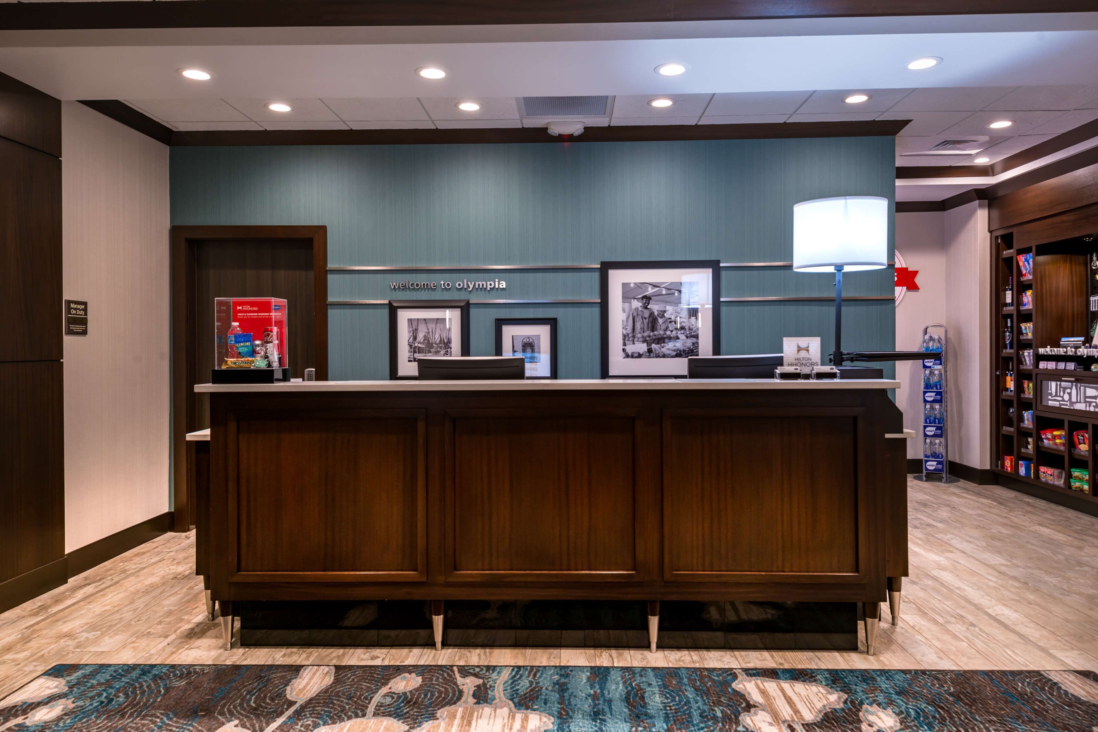 A reception desk with a sign that reads "Welcome to Olympia," framed pictures, a lamp, and a water dispenser.