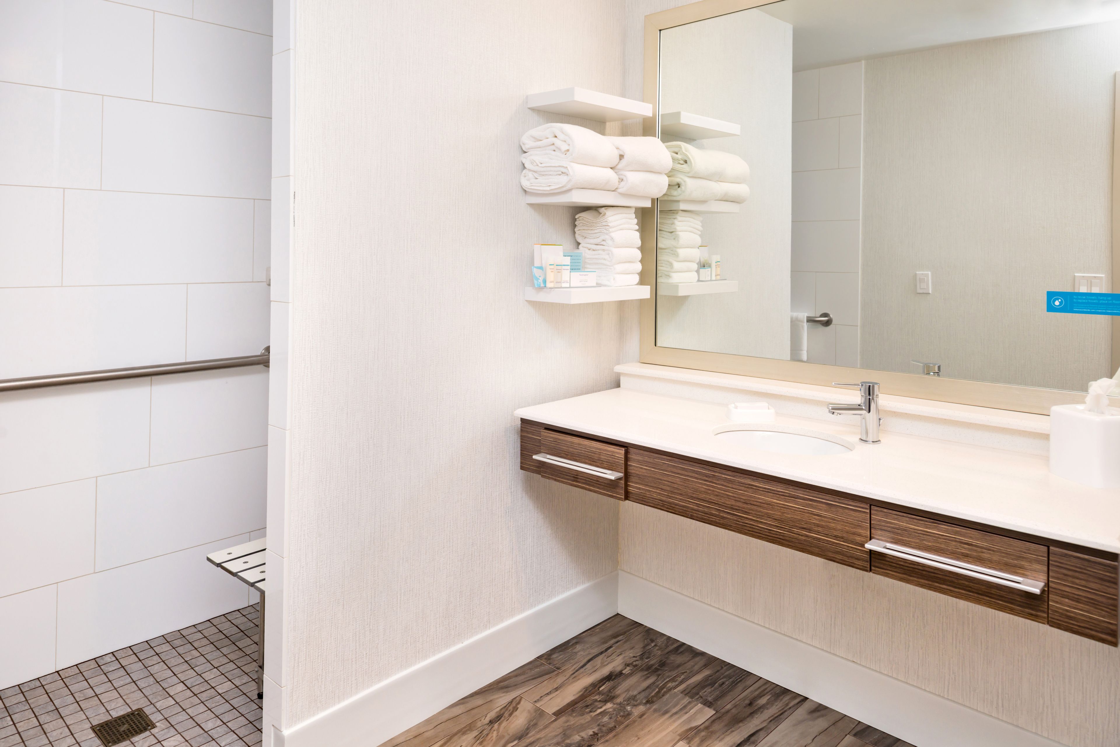 A modern bathroom with a wooden vanity, white tiles, a large mirror, and neatly folded towels on a shelf.