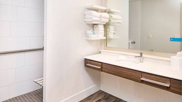 A modern bathroom with a wooden vanity, white tiles, a large mirror, and neatly folded towels on a shelf.