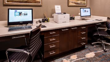 A hotel office with two desktop computers, a printer, and a keyboard. There are two chairs, a desk with drawers, and a carpeted floor.