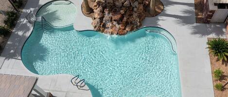 A large outdoor swimming pool with a waterfall feature, surrounded by lounge chairs and palm trees.