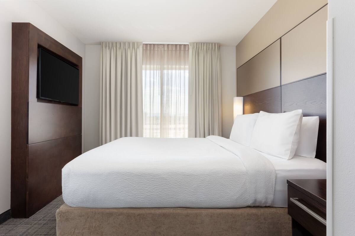 A hotel room with a large bed, a flat-screen TV mounted on the wall, and a window with curtains.