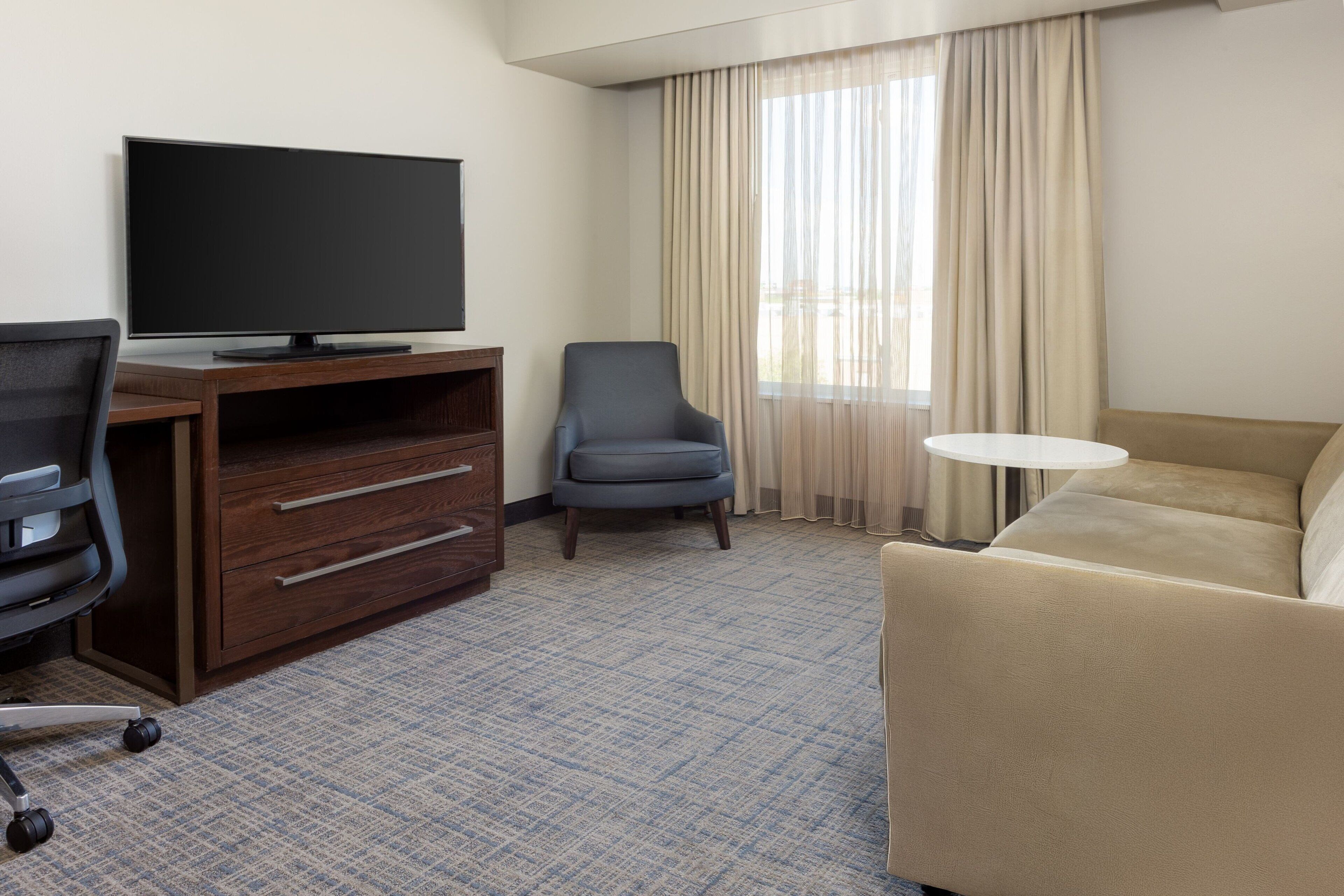 Habitación de hotel con televisión de pantalla plana, escritorio con cajones, una silla, un sofá y una mesita redonda.