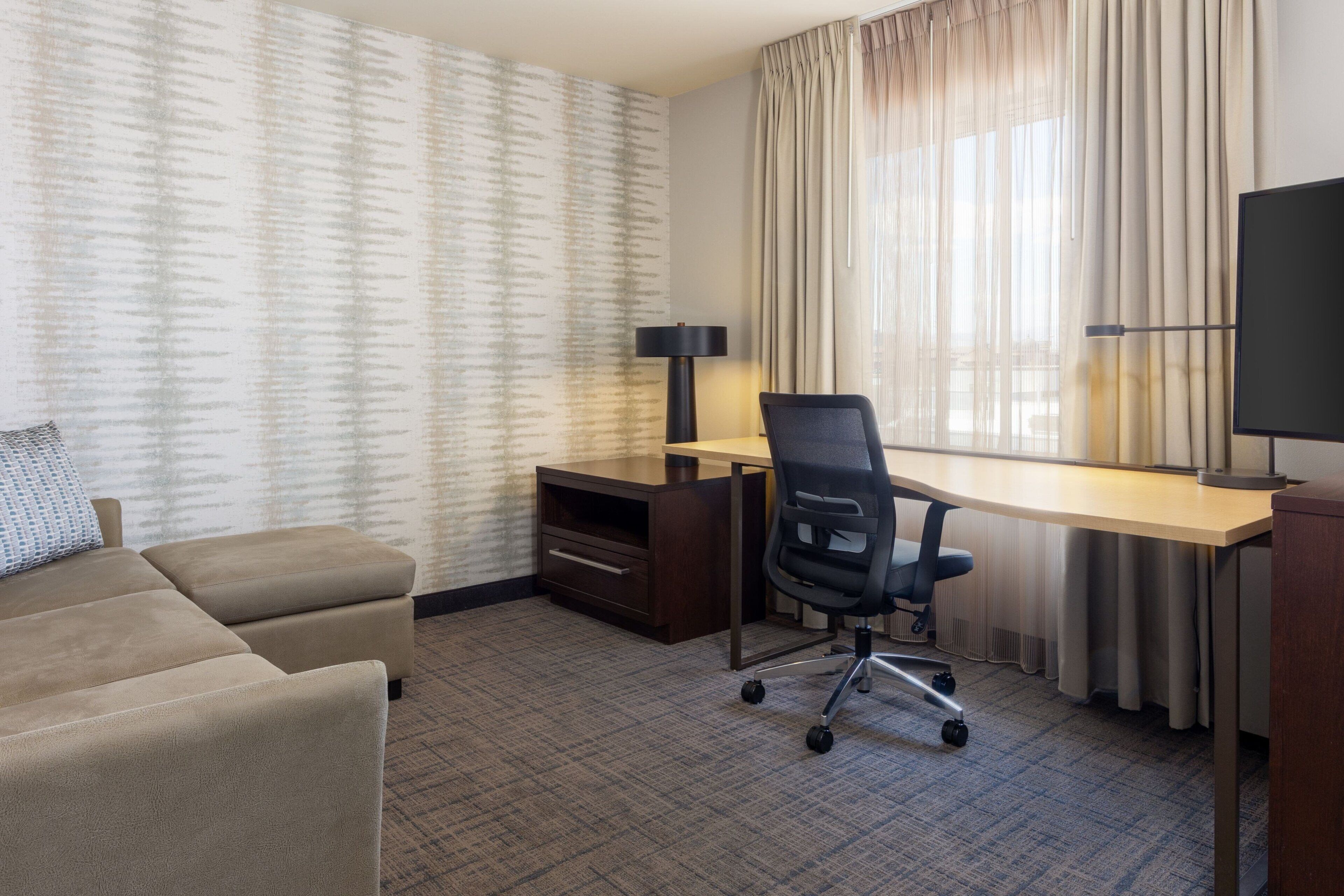 A hotel room with a sofa, a desk, a chair, a TV, and a window with curtains.