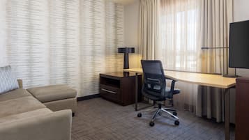 A hotel room with a sofa, a desk, a chair, a TV, and a window with curtains.