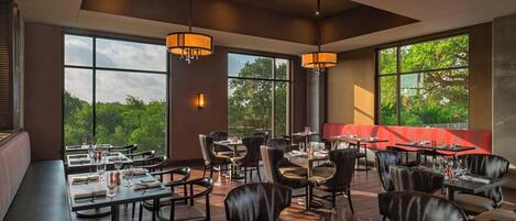A restaurant with high ceilings, large windows, and leather seating. Tables are set with wine glasses and cutlery.
