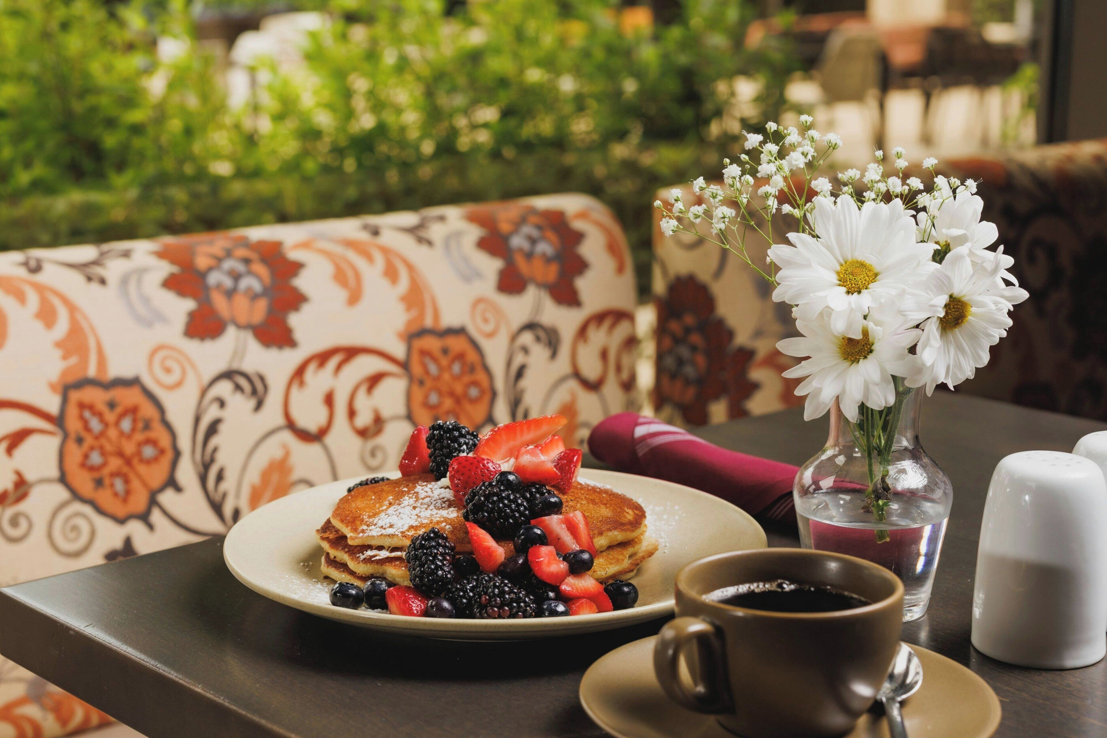A breakfast setting with pancakes, berries, a cup of coffee, and a vase of flowers on a table.