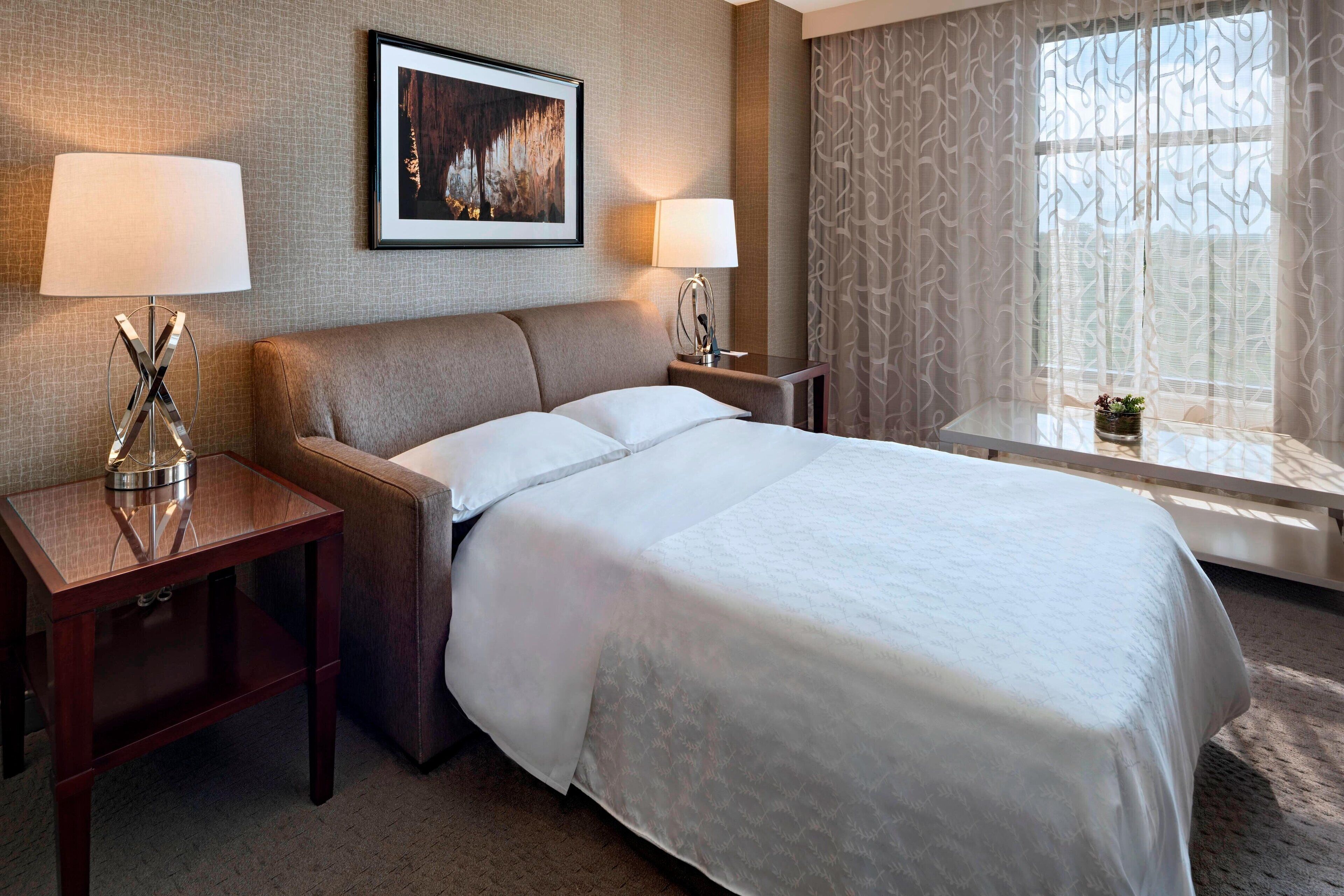 Une chambre d’hôtel avec un grand lit, deux tables de chevet avec des lampes, un tableau encadré au mur et une fenêtre avec des rideaux.