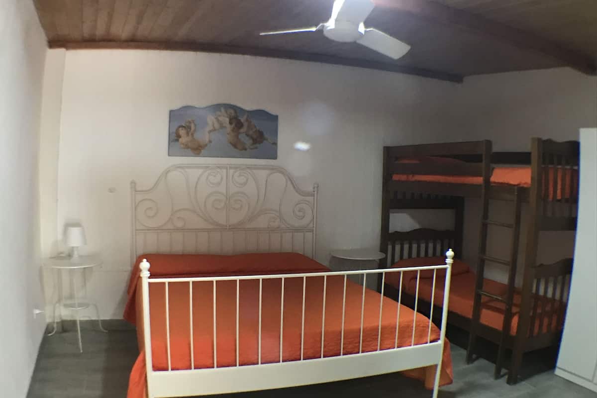 Ein Schlafzimmer mit einem weißen Metallbett, einem kleinen Tisch, einem Stuhl, einem Deckenventilator und einem Etagenbett.