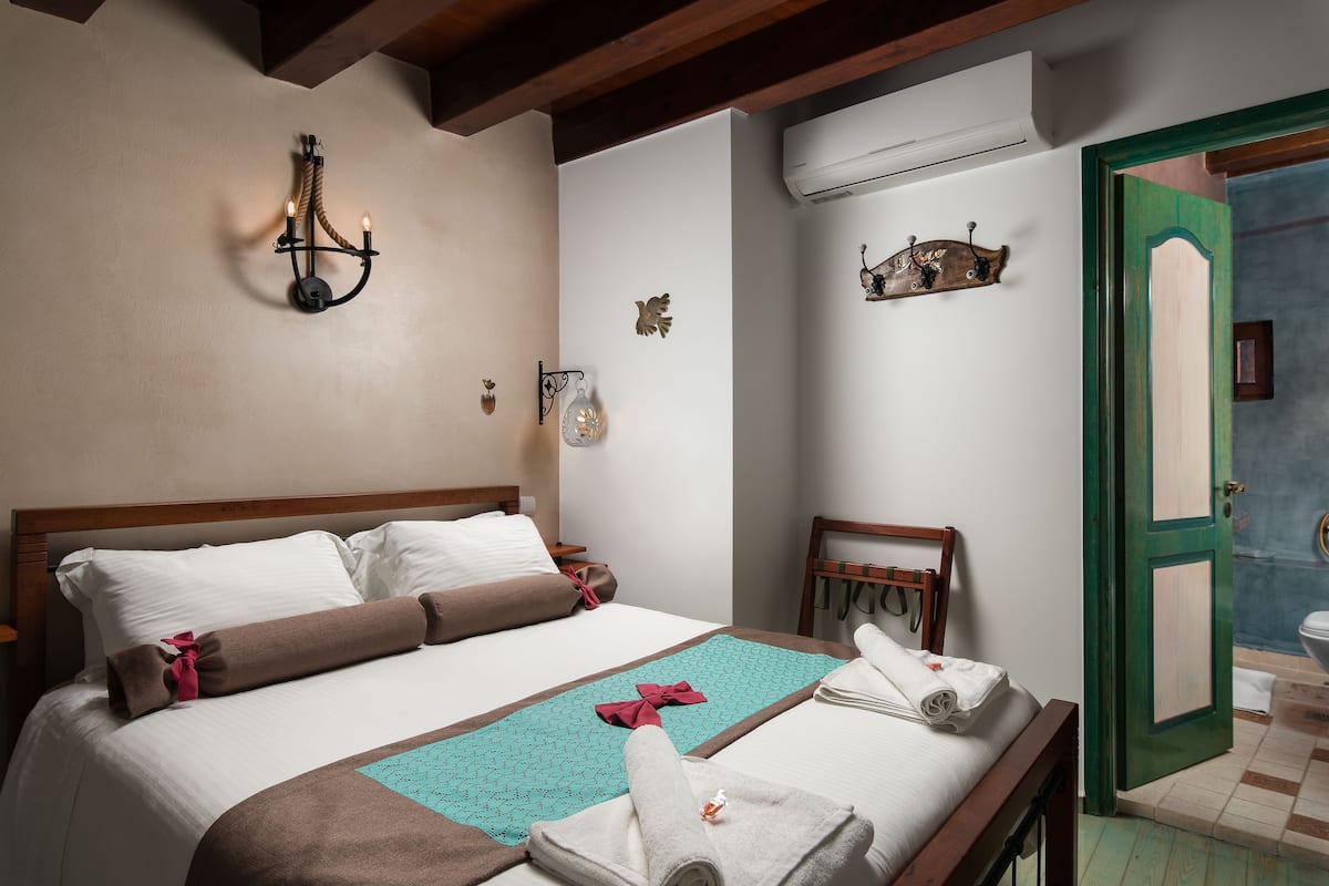 A neatly made bed with a teal blanket, white pillows, and a brown throw. There is a wooden headboard, a wall-mounted light fixture, and a small shelf with decorative items.