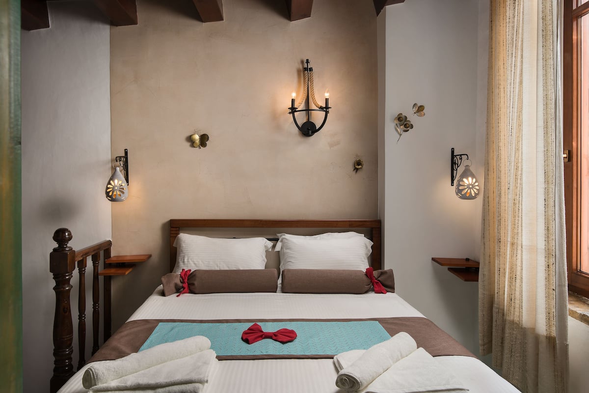 A neatly made bed with pillows, a decorative bow, and towels, a wooden headboard, and wall-mounted light fixtures.