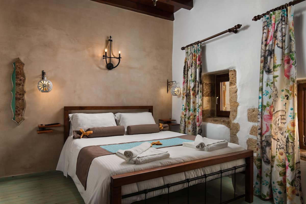A neatly made bed with a wooden headboard, a wall-mounted candelabra, and a floral curtain.
