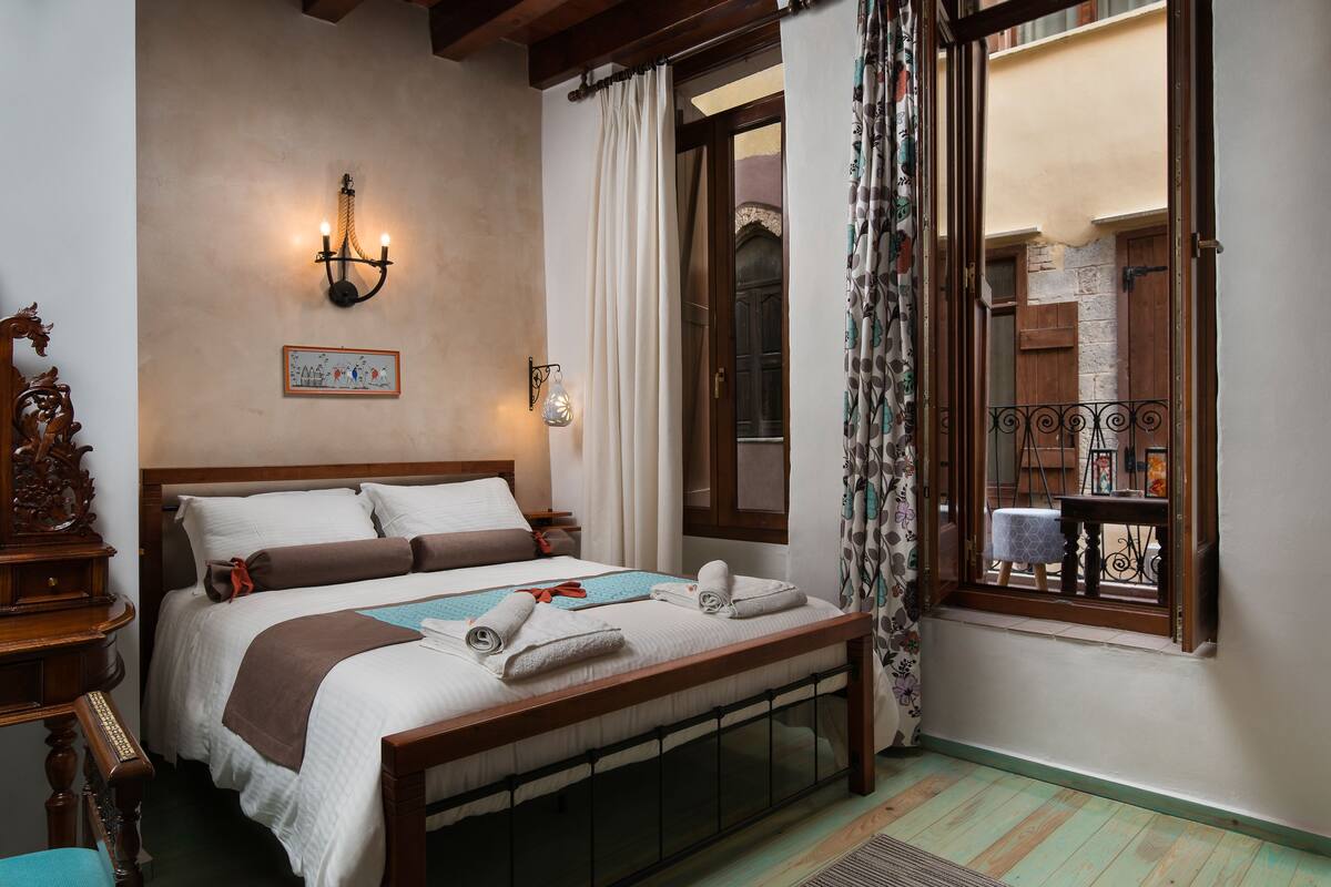 A bedroom with a wooden bed, a wall-mounted light, a window with curtains, and a view of the outside.