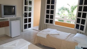 Habitación de hotel con cama, televisor, refrigerador pequeño y balcón con vista a follaje.