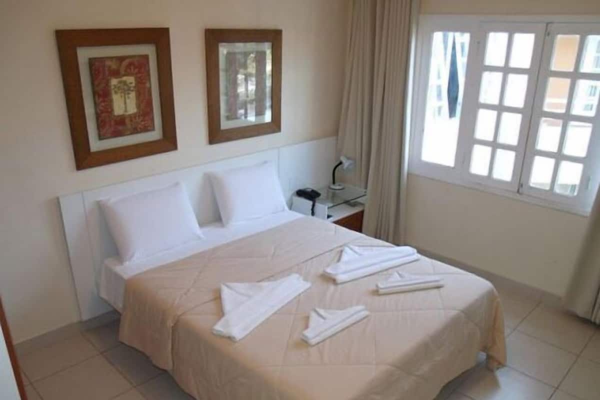 Habitación de hotel con cama, dos cuadros en la pared, una mesita de noche con lámpara y una ventana con cortinas.