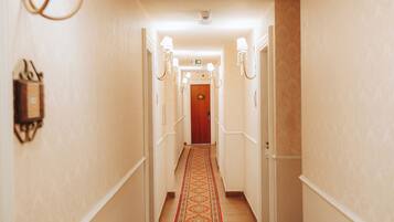 A hotel hallway with a patterned carpet, wall sconces, and a wooden door at the end.