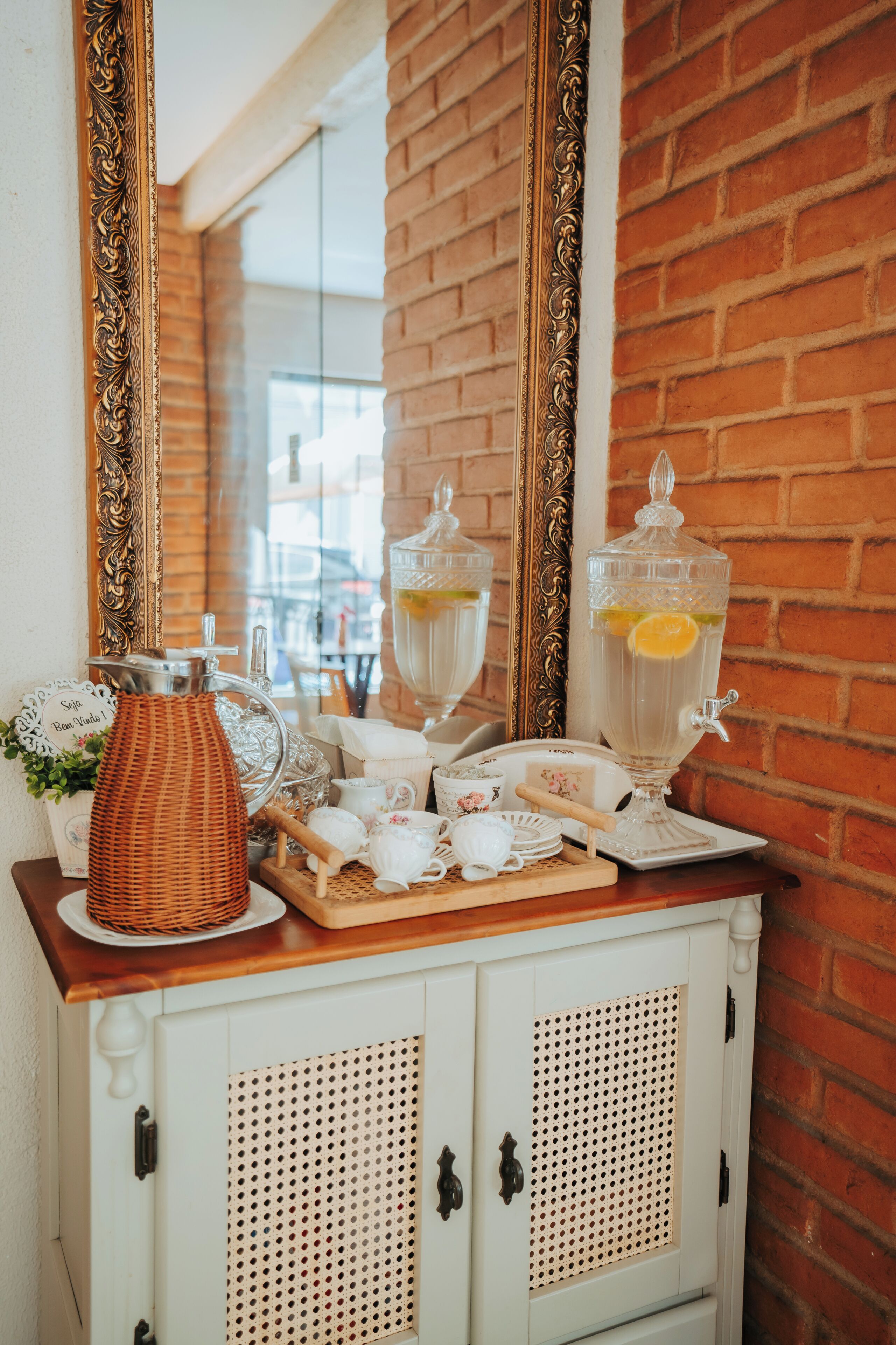 Una preparación para el desayuno que incluye una cesta de mimbre, tetera, tazas y un espejo que refleja un muro de ladrillo.
