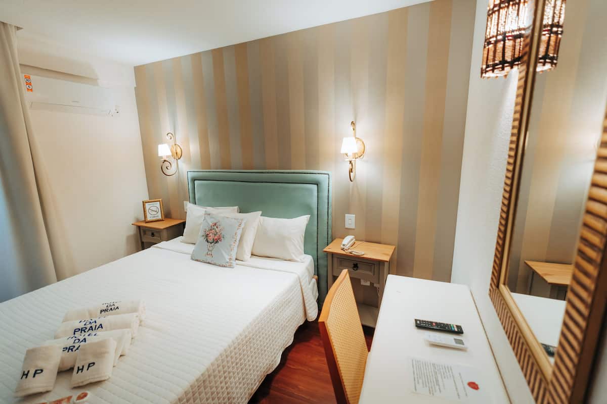A hotel room with a bed, bedside tables, a desk, a chair, a lamp, a mirror, and an air conditioning unit.