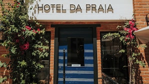 L’entrée d’un hôtel, avec une porte bleue, un balcon orné de balustrades en fer forgé et une enseigne indiquant « HÔTEL DA PRAIA ».