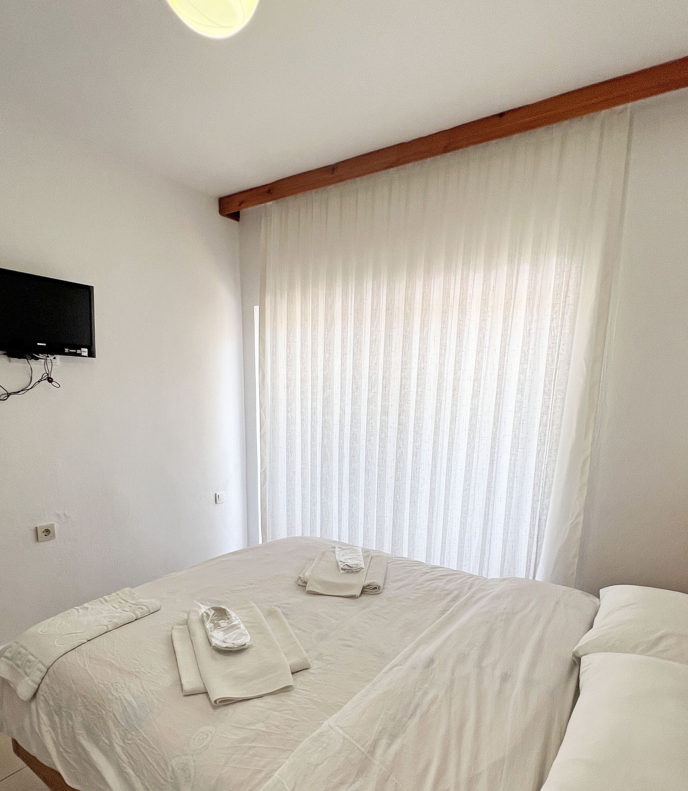 A room with two beds, a flat-screen TV mounted on the wall, and a window with sheer curtains.