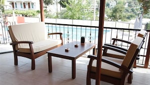 Un balcón con mobiliario de madera, que incluye dos sillas y una mesa, con vistas a una piscina y follaje.