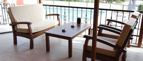 A balcony with wooden furniture, including two chairs and a table, overlooking a swimming pool and greenery.