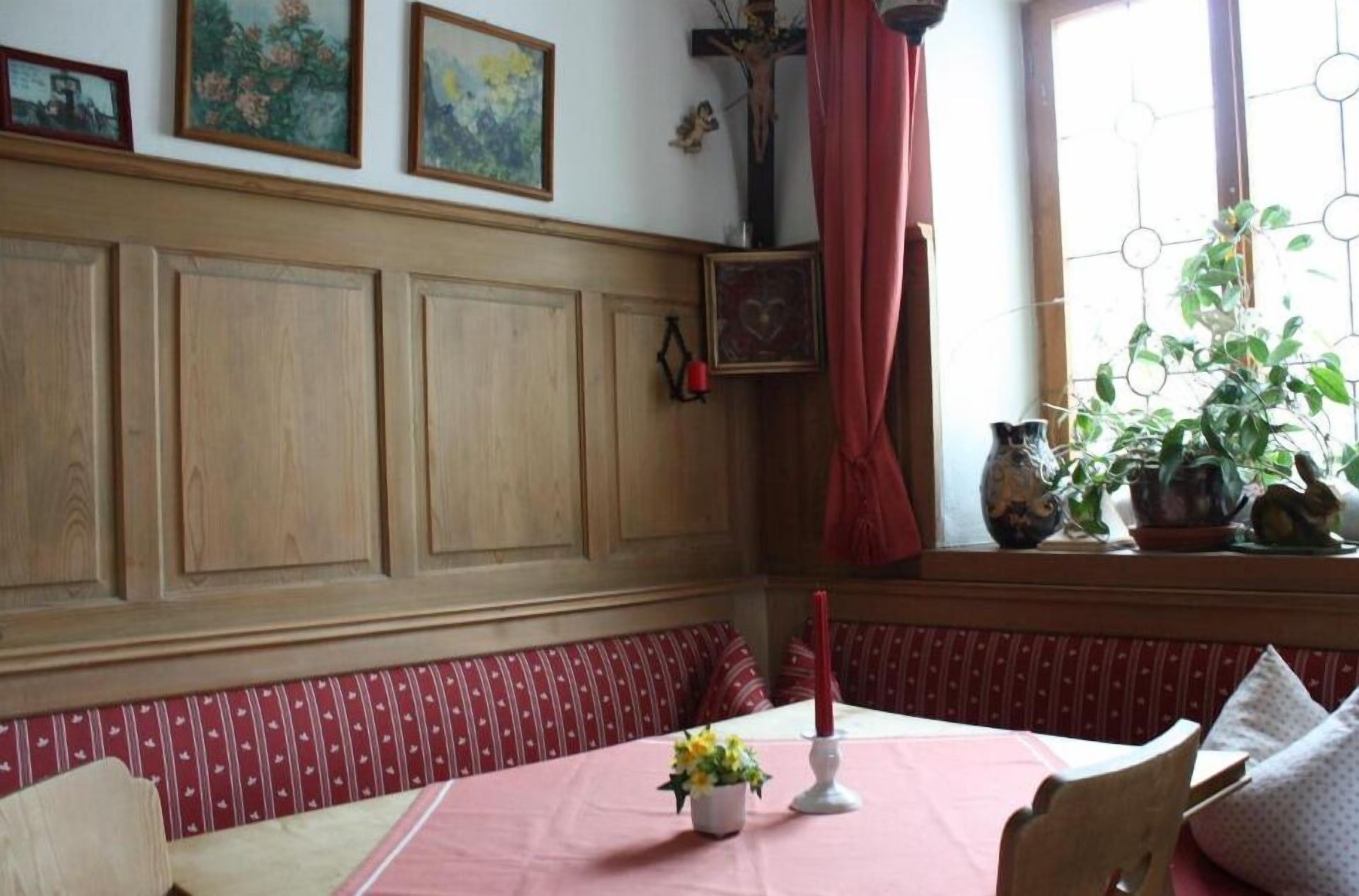 Une pièce avec un mur lambrissé en bois, un banc rouge, une table recouverte d’une nappe rose et une fenêtre ornée de plantes.