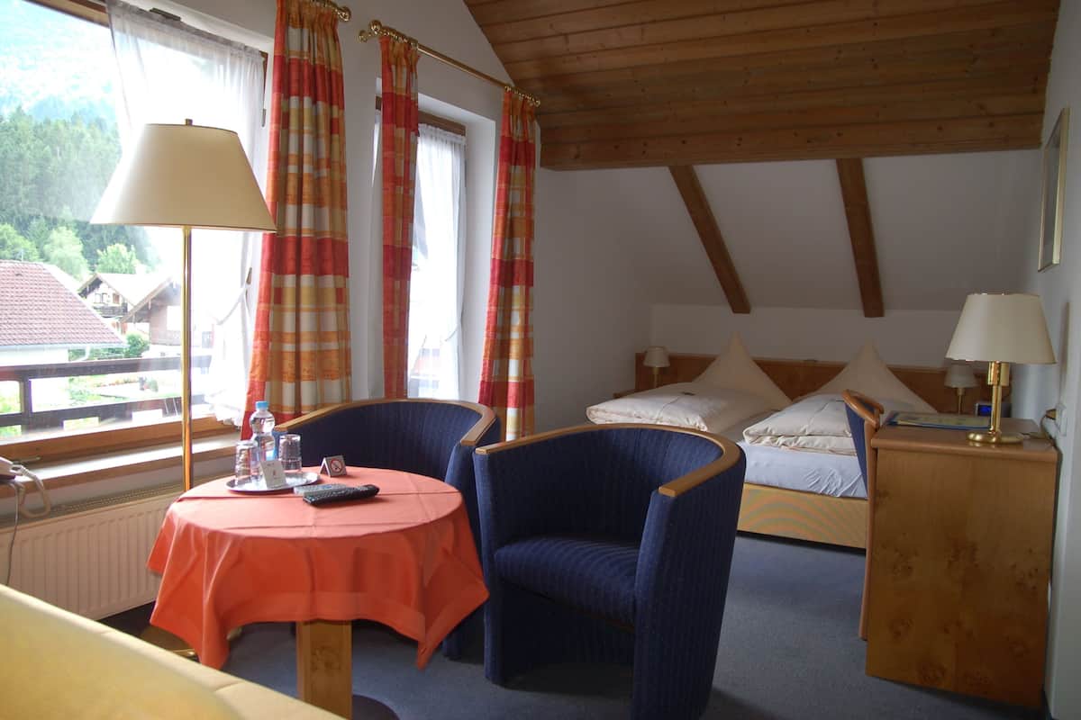 Ein Zimmer mit einem Bett, zwei Stühlen, einem Tisch mit Tischdecke, einer Lampe und Blick auf Häuser.