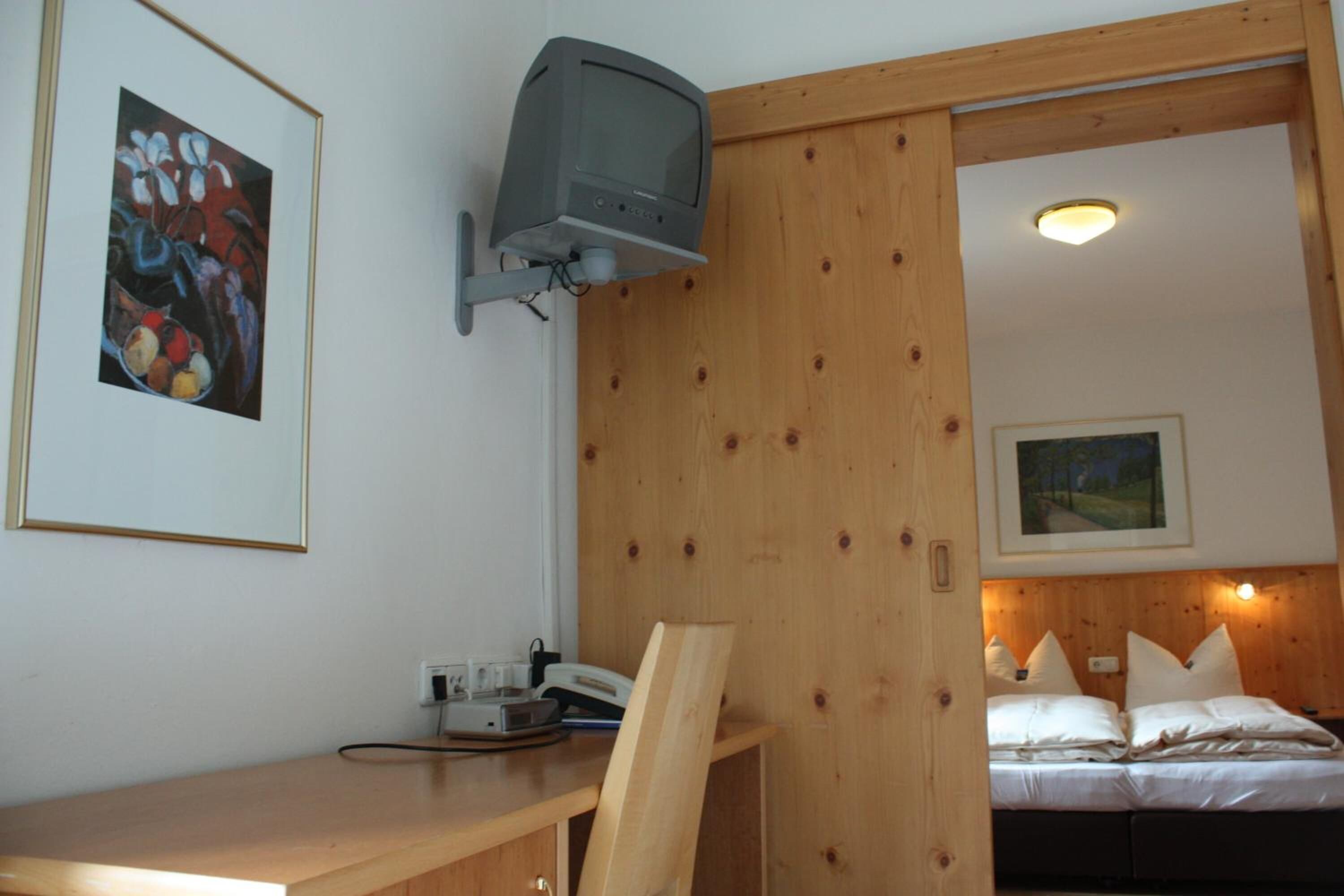Ein Zimmer mit einem Holztisch, einem an der Wand montierten Fernseher, einem Bett mit weißer Bettwäsche und einem gerahmten Gemälde an der Wand.