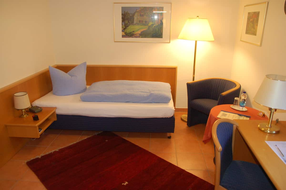 Ein Hotelzimmer mit Bett, Schreibtisch, Stuhl, Lampe, Nachttisch und Teppich.