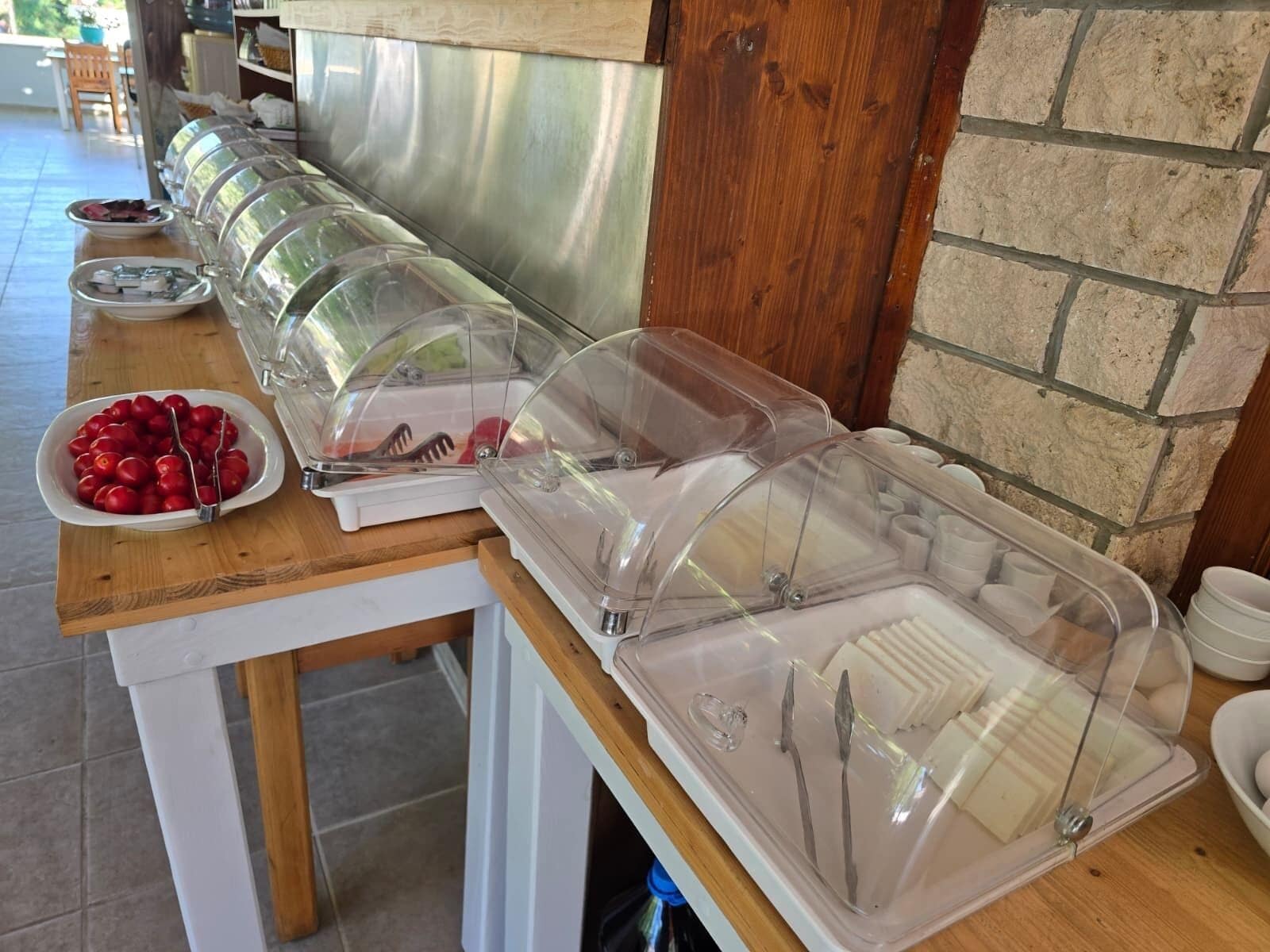 A buffet setup with clear plastic covers over food containers, a bowl of cherries, and a brick wall in the background.