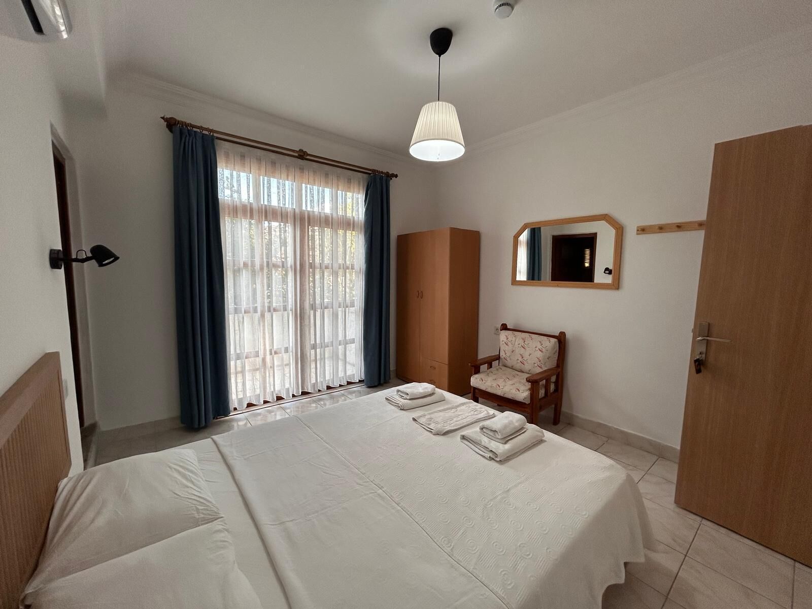 A hotel room with a bed, a chair, a wardrobe, a mirror, and a window with curtains.