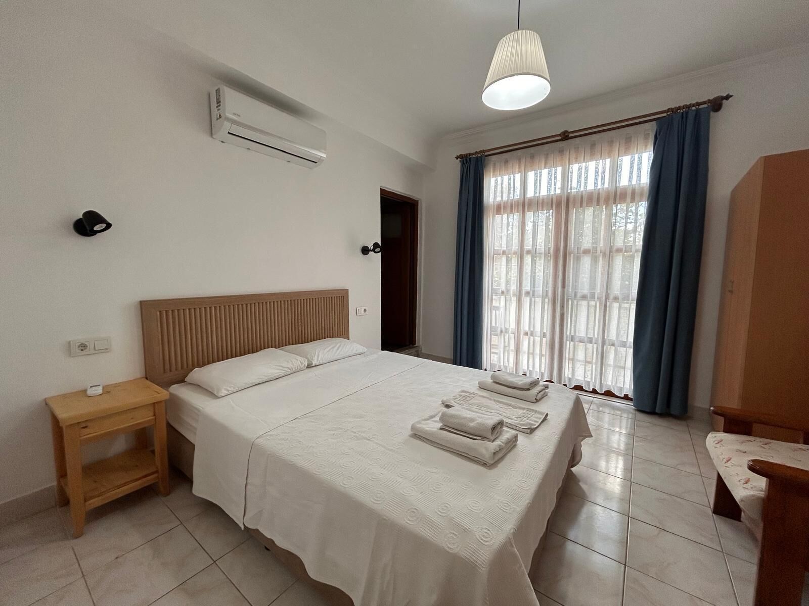 Ein Schlafzimmer mit Bett, Nachttisch, Klimaanlage, einem Fenster mit Vorhängen und einer Pendelleuchte.