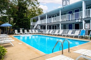 A swimming pool with white lounge chairs and a blue umbrella, surrounded by a two-story building with balconies.