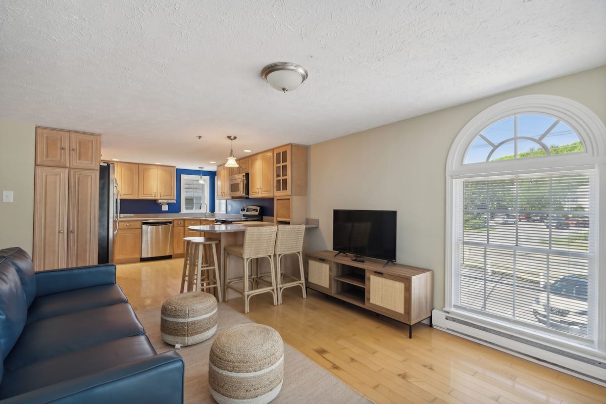 A modern kitchen with wooden cabinets, a dining area with a bar stool, a flat-screen TV, and a large window with blinds.