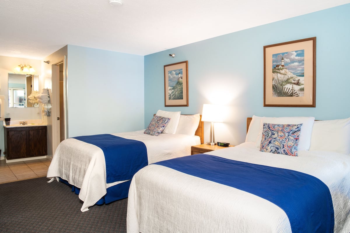 A hotel room with two beds, a nightstand with a lamp, a dresser, and a mirror.