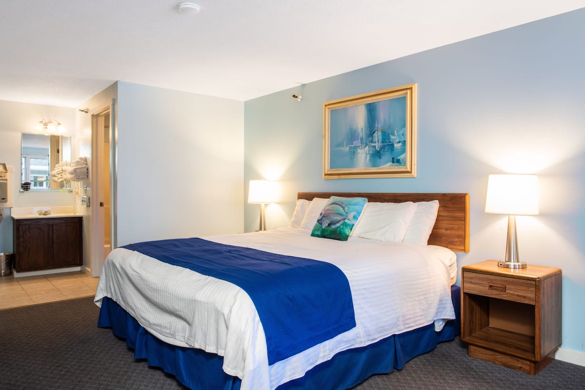 A hotel room with a large bed, two bedside lamps, a wooden nightstand, a painting on the wall, and a bathroom visible through an open door.